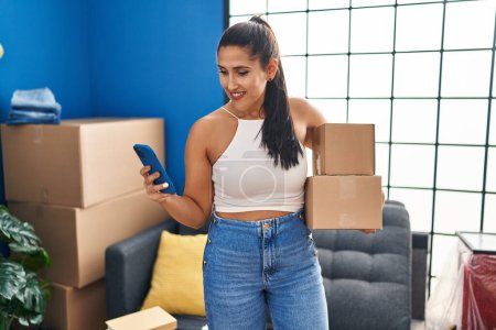 Photo for Young hispanic woman using smartphone holding packages at new home - Royalty Free Image