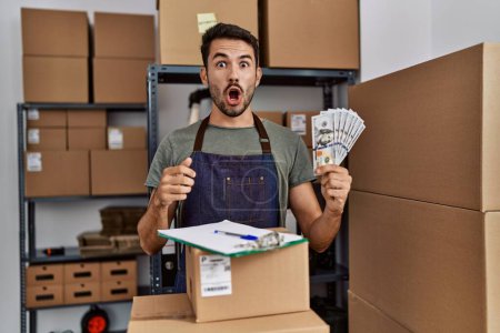 Photo for Young hispanic man working at small business ecommerce holding money scared and amazed with open mouth for surprise, disbelief face - Royalty Free Image