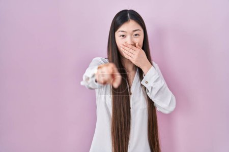 Foto de Mujer joven china de pie sobre fondo rosa riéndose de ti, señalando con el dedo a la cámara con la mano sobre la boca, expresión de vergüenza - Imagen libre de derechos