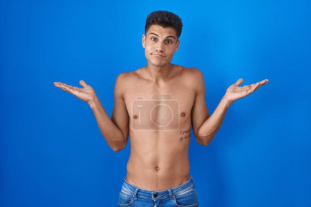 Photo for Young hispanic man standing shirtless over blue background clueless and confused expression with arms and hands raised. doubt concept. - Royalty Free Image