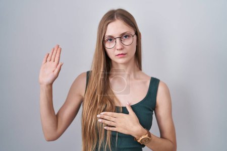 Foto de Mujer caucásica joven de pie sobre fondo blanco jurando con la mano en el pecho y la palma abierta, haciendo una promesa de lealtad juramento - Imagen libre de derechos