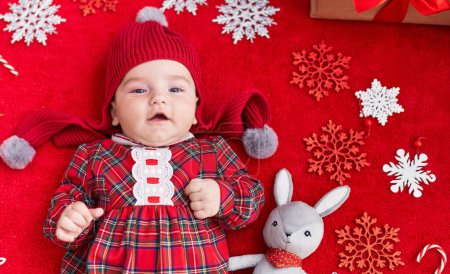 Téléchargez les photos : Adorable bébé caucasien couché sur le sol avec décor de Noël sur fond rouge isolé - en image libre de droit