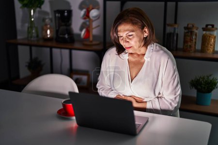 Téléchargez les photos : Femme hispanique du moyen âge utilisant un ordinateur portable à la maison la nuit avec la main sur le ventre parce que l'indigestion, la maladie douloureuse se sent mal. concept de douleur. - en image libre de droit