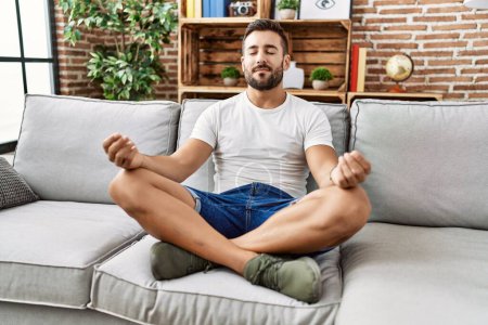 Photo for Young hispanic man doing yoga exercise sitting on the sofa at home - Royalty Free Image
