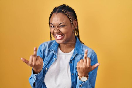 Photo for African american woman with braids standing over yellow background showing middle finger doing fuck you bad expression, provocation and rude attitude. screaming excited - Royalty Free Image