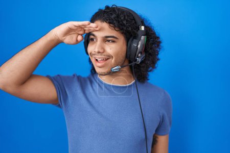 Photo for Hispanic man with curly hair listening to music using headphones very happy and smiling looking far away with hand over head. searching concept. - Royalty Free Image