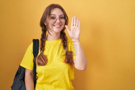 Foto de Joven mujer caucásica usando mochila de estudiante sobre fondo amarillo renunciando a decir hola feliz y sonriente, gesto de bienvenida amistoso - Imagen libre de derechos