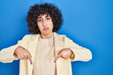 Photo for Young brunette woman with curly hair standing over blue background pointing down looking sad and upset, indicating direction with fingers, unhappy and depressed. - Royalty Free Image