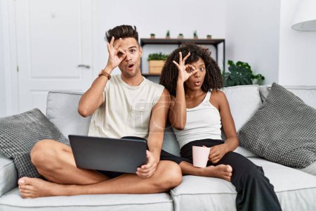 Téléchargez les photos : Jeune couple interracial utilisant un ordinateur portable à la maison assis sur le canapé faisant un geste correct choqué par le visage surpris, les yeux regardant à travers les doigts. expression incrédule. - en image libre de droit