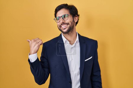 Foto de Handsome latin man standing over yellow background smiling with happy face looking and pointing to the side with thumb up. - Imagen libre de derechos