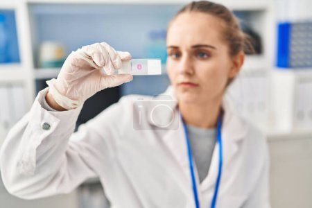 Téléchargez les photos : Jeune femme blonde portant l'uniforme de scientifique regardant l'échantillon au laboratoire - en image libre de droit
