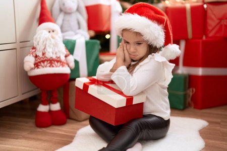 Téléchargez les photos : Adorable fille hispanique tenant cadeau assis sur le sol par l'arbre de Noël avec une expression triste à la maison - en image libre de droit