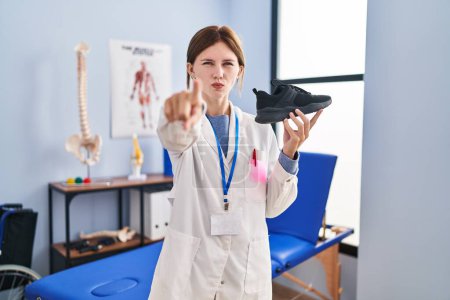 Téléchargez les photos : Jeune physiothérapeute femme tenant des baskets pointant du doigt vers la caméra et vers vous, geste confiant et sérieux - en image libre de droit