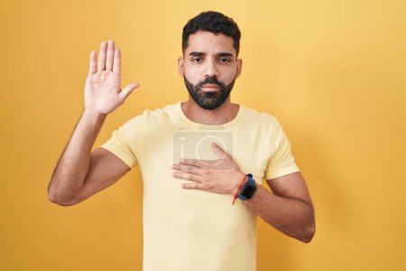 Photo for Hispanic man with beard standing over yellow background swearing with hand on chest and open palm, making a loyalty promise oath - Royalty Free Image