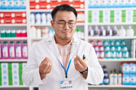Téléchargez les photos : Jeune homme chinois travaillant à la pharmacie pharmacie faire un geste d'argent avec les mains, demander le paiement du salaire, millionnaire entreprise - en image libre de droit