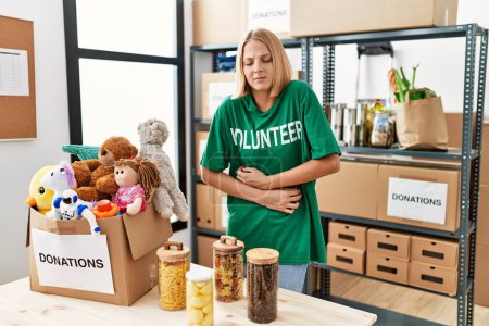 Foto de Young caucasian woman wearing volunteer t shirt at donations stand with hand on stomach because nausea, painful disease feeling unwell. ache concept. - Imagen libre de derechos