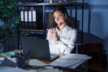 Photo for Young brunette woman wearing call center agent headset working late at night cheerful with a smile of face pointing with hand and finger up to the side with happy and natural expression on face - Royalty Free Image