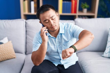 Foto de Young chinese man talking on the smartphone looking watch at home - Imagen libre de derechos