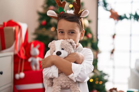 Téléchargez les photos : Adorable bambin hispanique étreignant ours en peluche debout près de l'arbre de Noël à la maison - en image libre de droit