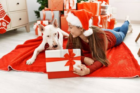 Photo for Young caucasian woman unboxing gift lying with dog by christmas tree at home - Royalty Free Image