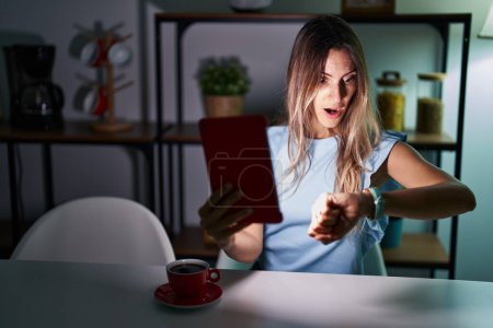 Foto de Mujer hispana joven usando touchpad sentada en la mesa por la noche mirando el reloj preocupado, con miedo de llegar tarde - Imagen libre de derechos