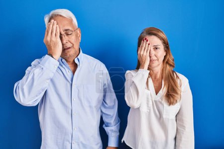 Photo for Middle age hispanic couple standing over blue background yawning tired covering half face, eye and mouth with hand. face hurts in pain. - Royalty Free Image