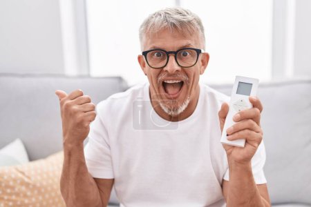 Téléchargez les photos : Homme hispanique aux cheveux gris tenant le contrôle du climatiseur pointant le pouce vers le côté souriant heureux avec la bouche ouverte - en image libre de droit