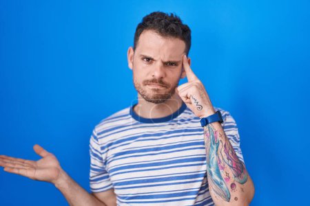 Téléchargez les photos : Young hispanic man standing over blue background confused and annoyed with open palm showing copy space and pointing finger to forehead. think about it. - en image libre de droit