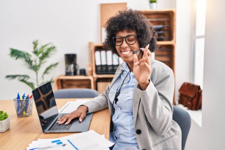 Foto de Black woman with curly hair wearing call center agent headset at the office gesturing finger crossed smiling with hope and eyes closed. luck and superstitious concept. - Imagen libre de derechos