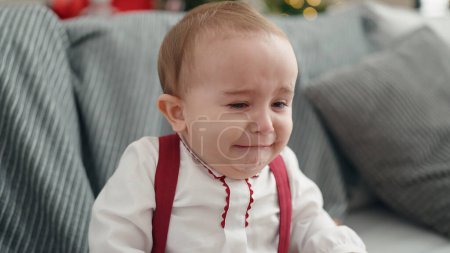 Téléchargez les photos : Adorable hispanic baby sitting on sofa by christmas tree crying at home - en image libre de droit