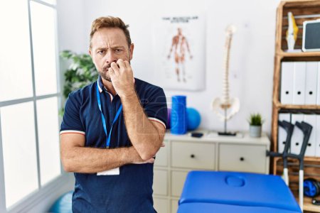 Photo for Middle age physiotherapist man working at pain recovery clinic looking stressed and nervous with hands on mouth biting nails. anxiety problem. - Royalty Free Image