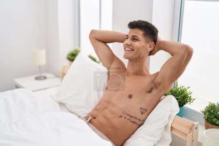 Téléchargez les photos : Jeune homme hispanique détendu avec les mains sur la tête assise sur le lit dans la chambre - en image libre de droit