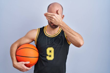 Téléchargez les photos : Jeune homme chauve avec barbe portant un uniforme de basket-ball tenant le ballon couvrant les yeux avec la main, l'air sérieux et triste. concept de non-visibilité, de dissimulation et de rejet - en image libre de droit