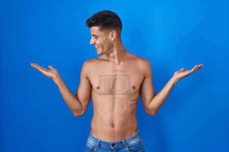 Photo for Young hispanic man standing shirtless over blue background smiling showing both hands open palms, presenting and advertising comparison and balance - Royalty Free Image