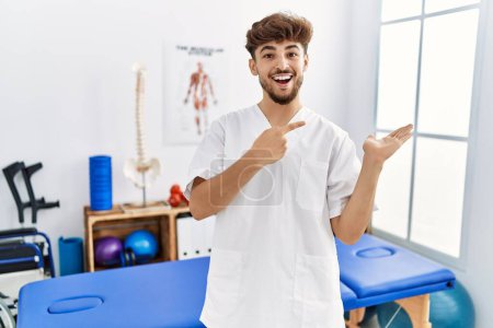 Photo for Young arab man working at pain recovery clinic amazed and smiling to the camera while presenting with hand and pointing with finger. - Royalty Free Image