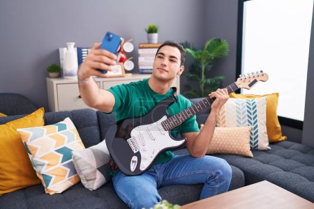 Foto de Hombre hispano joven tocando la guitarra eléctrica hacer selfie por teléfono inteligente en casa - Imagen libre de derechos