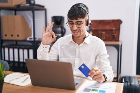 Foto de Joven hombre hispano que trabaja usando computadora portátil con tarjeta de crédito sonriendo positiva haciendo signo de ok con la mano y los dedos. expresión exitosa. - Imagen libre de derechos
