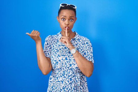 Foto de Mujer afroamericana de pie sobre fondo azul pidiendo estar callada con el dedo en los labios apuntando con la mano hacia un lado. silencio y concepto secreto. - Imagen libre de derechos