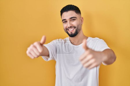 Photo for Young handsome man wearing casual t shirt over yellow background approving doing positive gesture with hand, thumbs up smiling and happy for success. winner gesture. - Royalty Free Image