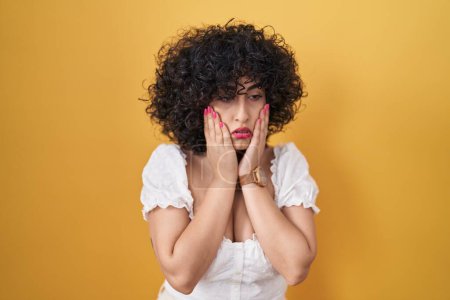 Foto de Young brunette woman with curly hair standing over yellow background tired hands covering face, depression and sadness, upset and irritated for problem - Imagen libre de derechos