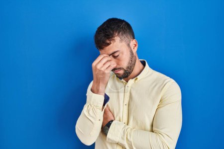 Foto de Handsome hispanic man standing over blue background tired rubbing nose and eyes feeling fatigue and headache. stress and frustration concept. - Imagen libre de derechos