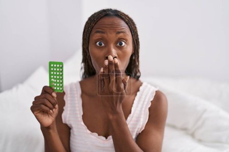 Téléchargez les photos : Femme afro-américaine tenant des pilules contraceptives couvrant la bouche avec la main, choquée et effrayée par l'erreur. expression surprise - en image libre de droit