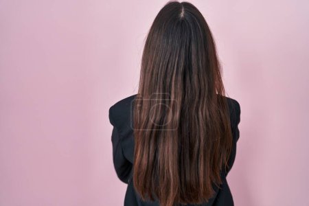 Téléchargez les photos : Jeune femme brune portant un style d'affaires sur fond rose debout vers l'arrière regardant loin avec les bras croisés - en image libre de droit
