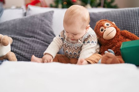 Photo for Adorable caucasian baby sitting on sofa by christmas tree with relaxed expression at home - Royalty Free Image