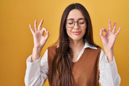 Téléchargez les photos : Jeune femme brune debout sur fond jaune portant des lunettes se détendre et sourire les yeux fermés en faisant un geste de méditation avec les doigts. concept de yoga. - en image libre de droit