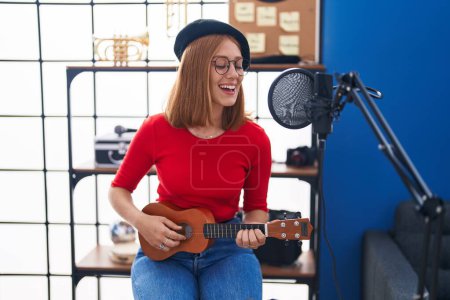 Téléchargez les photos : Jeune rousse femme musicienne chantant chanson jouant ukulele au studio de musique - en image libre de droit