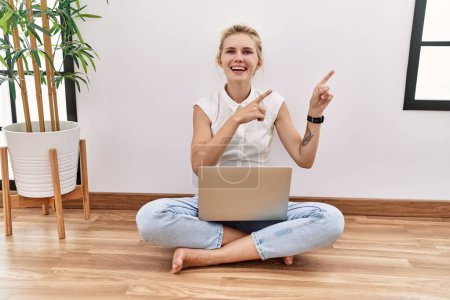 Foto de Mujer rubia joven usando computadora portátil sentada en el suelo en la sala de estar sonriendo y mirando a la cámara apuntando con dos manos y dedos hacia un lado. - Imagen libre de derechos