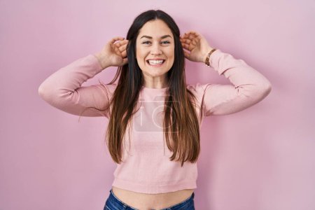 Foto de Mujer morena joven de pie sobre fondo rosa sonriendo tirando de las orejas con los dedos, gesto divertido. problema de audición - Imagen libre de derechos