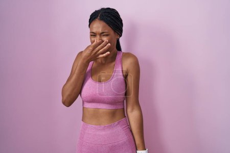 Foto de African american woman with braids wearing sportswear over pink background smelling something stinky and disgusting, intolerable smell, holding breath with fingers on nose. bad smell - Imagen libre de derechos
