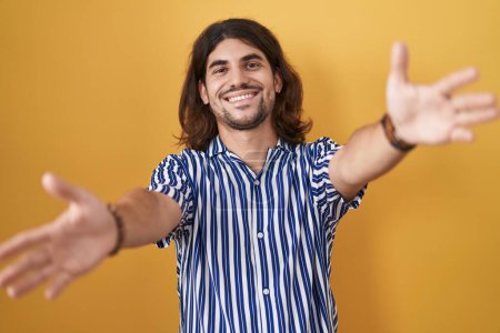 Téléchargez les photos : Homme hispanique avec les cheveux longs debout sur fond jaune regardant la caméra souriant à bras ouverts pour étreindre. expression joyeuse embrassant le bonheur. - en image libre de droit